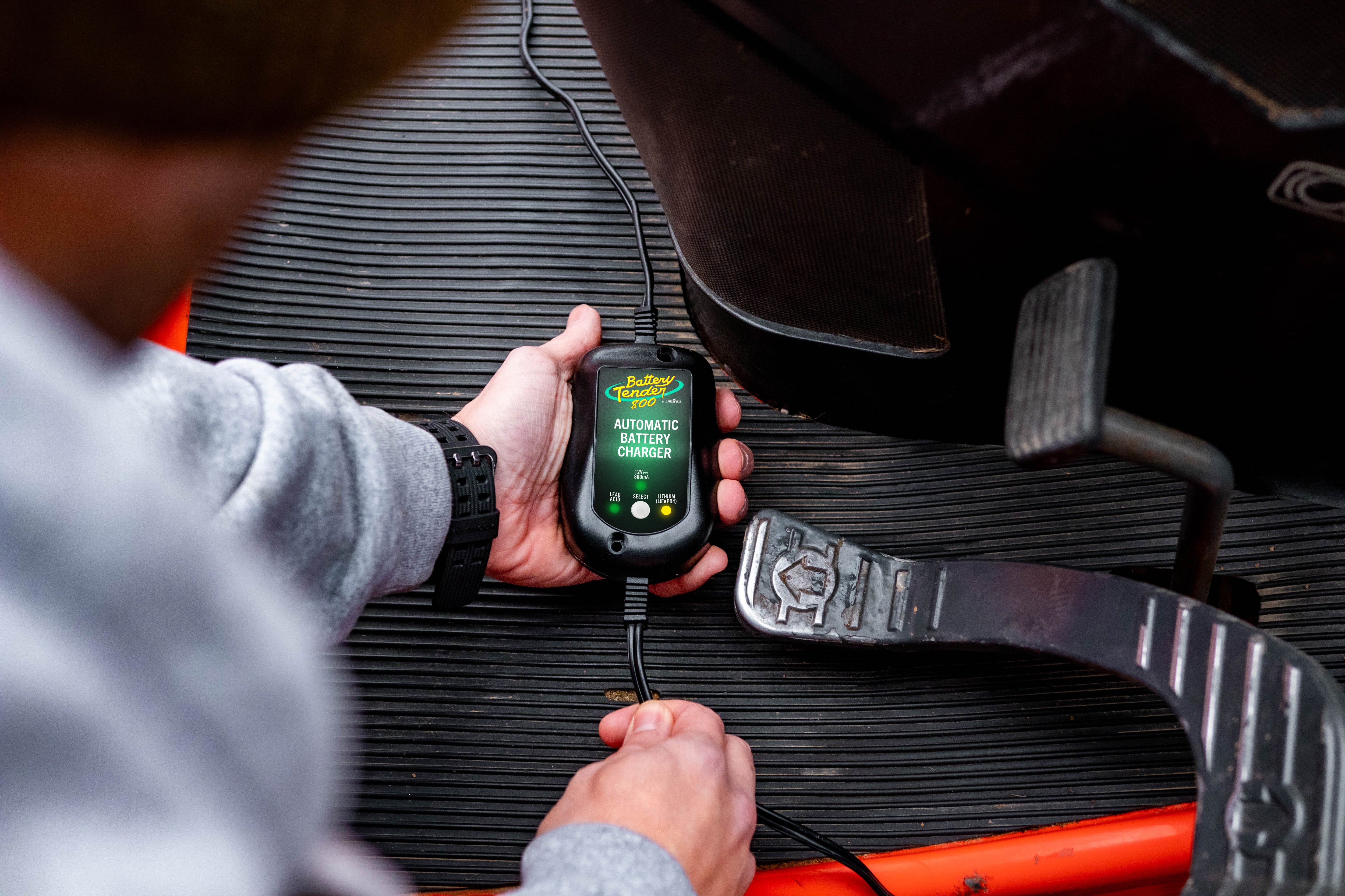 A man holds a selectable battery tender charger in his hand after connecting it to his vehicle's battery.